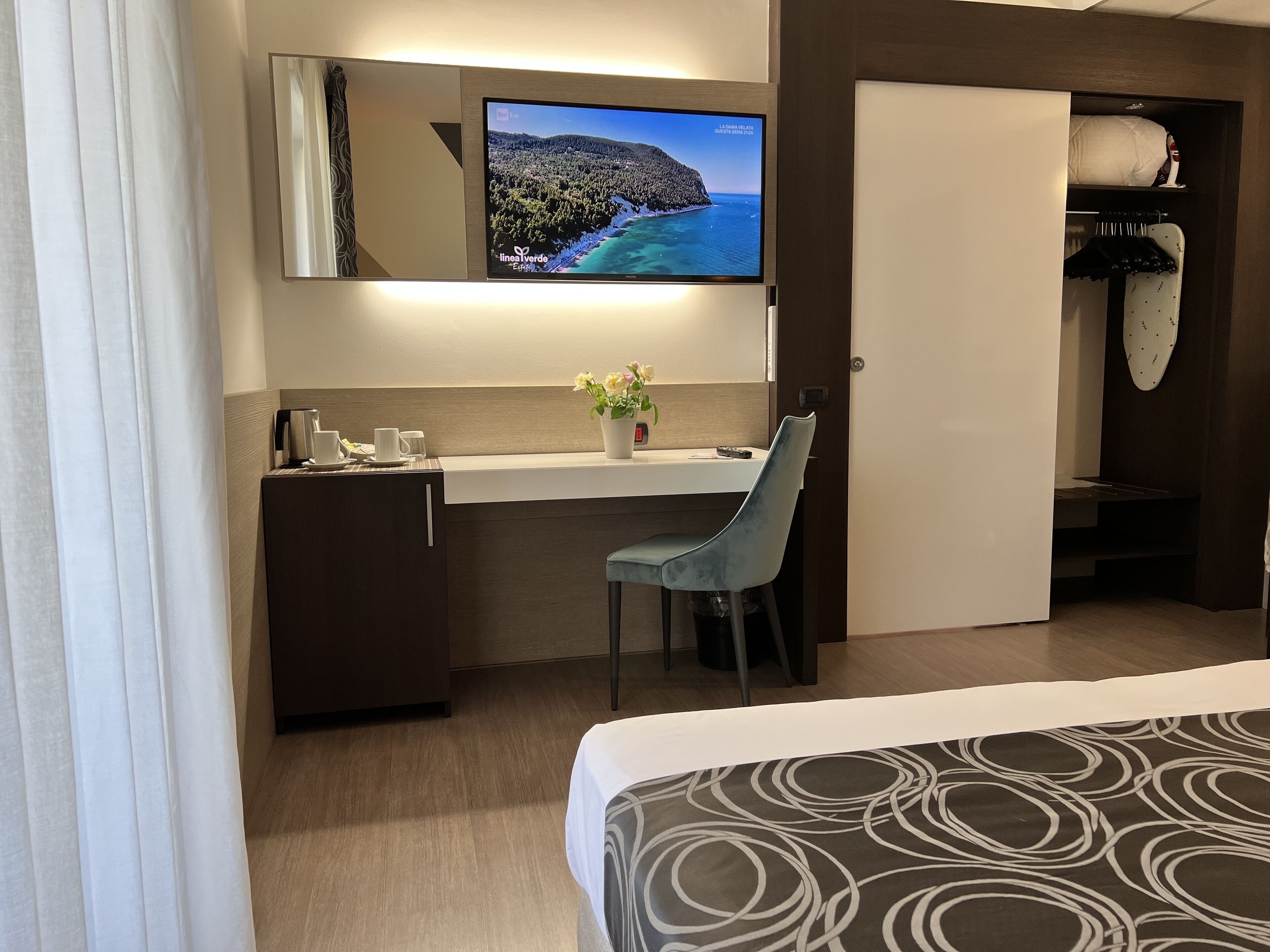 Una habitación de hotel con cama, escritorio con silla, televisor montado en la pared, armario y ventana con cortinas.