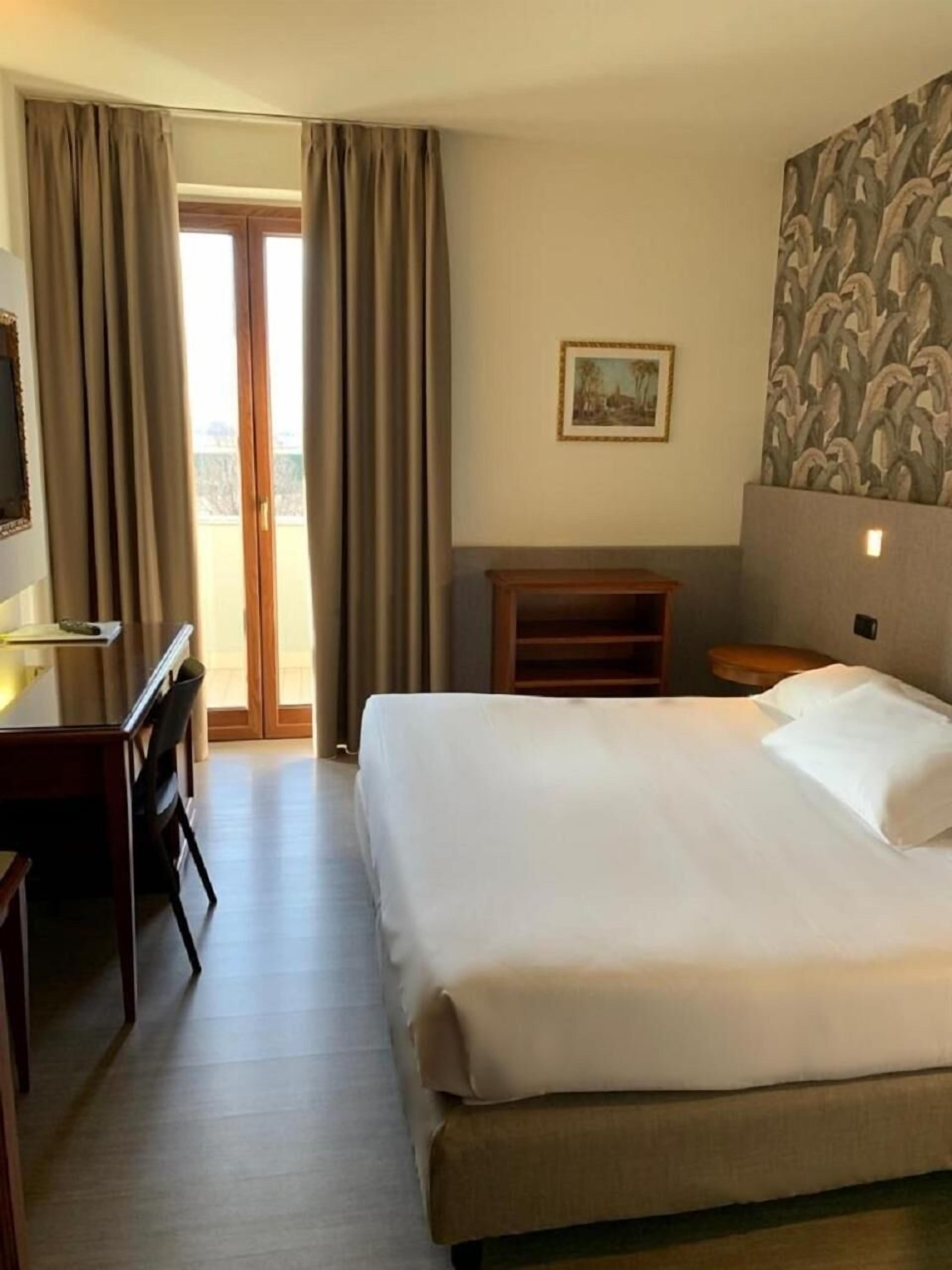 A hotel room with a bed, a desk, a chair, a small table, a picture on the wall, and a view through the door.