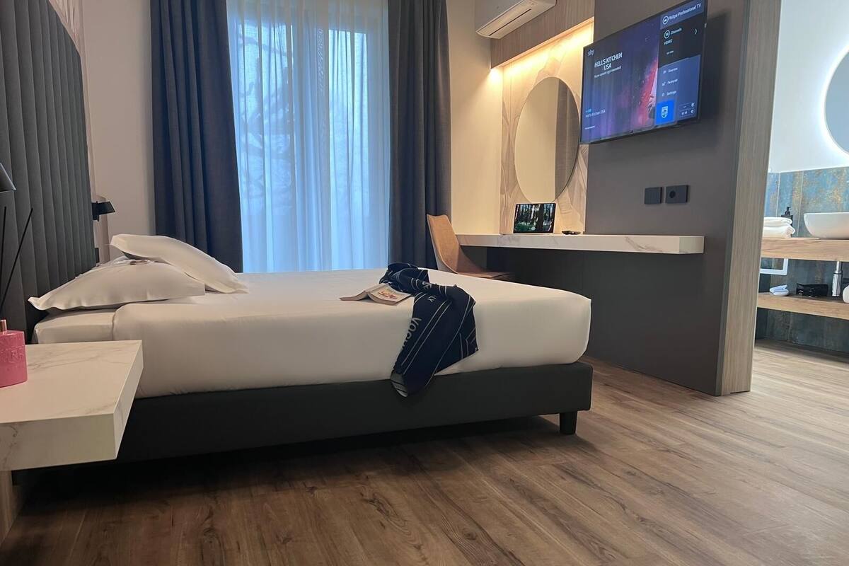 Une chambre d’hôtel moderne avec un grand lit, une table de chevet avec une lampe, un téléviseur fixé au mur et une petite salle de bain séparée par une cloison en verre.