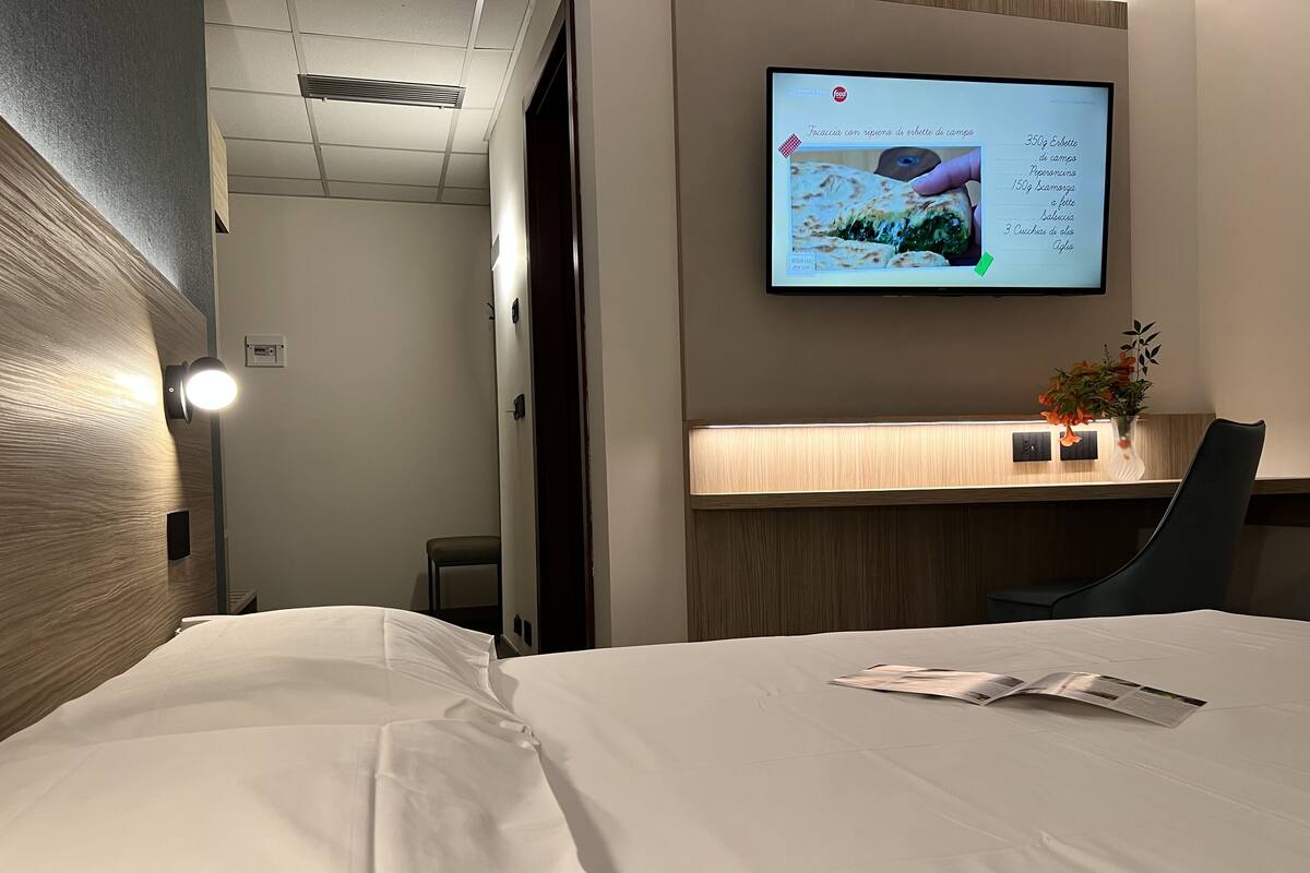Une chambre d’hôtel avec un lit, un bureau doté d’un écran d’ordinateur affichant un menu, une chaise et une petite table sur laquelle est posé un vase de fleurs.