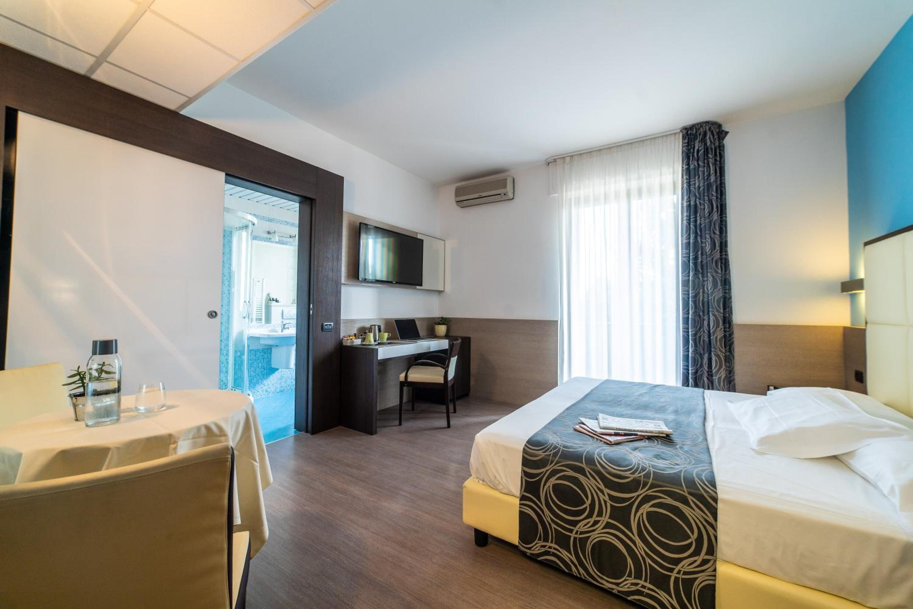 Habitación de hotel con cama, escritorio, silla, televisión y ventana con cortinas.