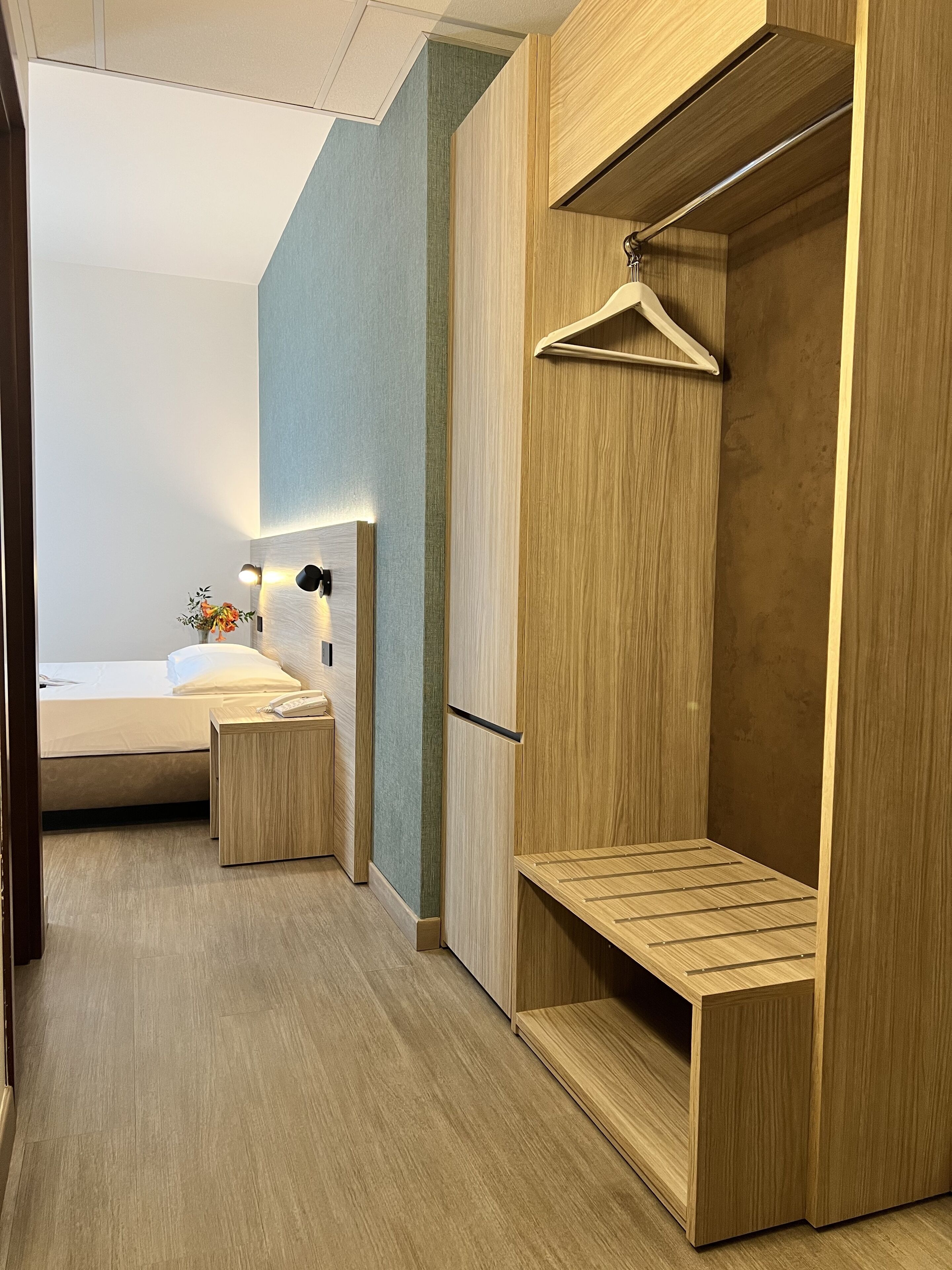 A modern hotel room with a bed, bedside table, and a wooden wardrobe with a hanging space.