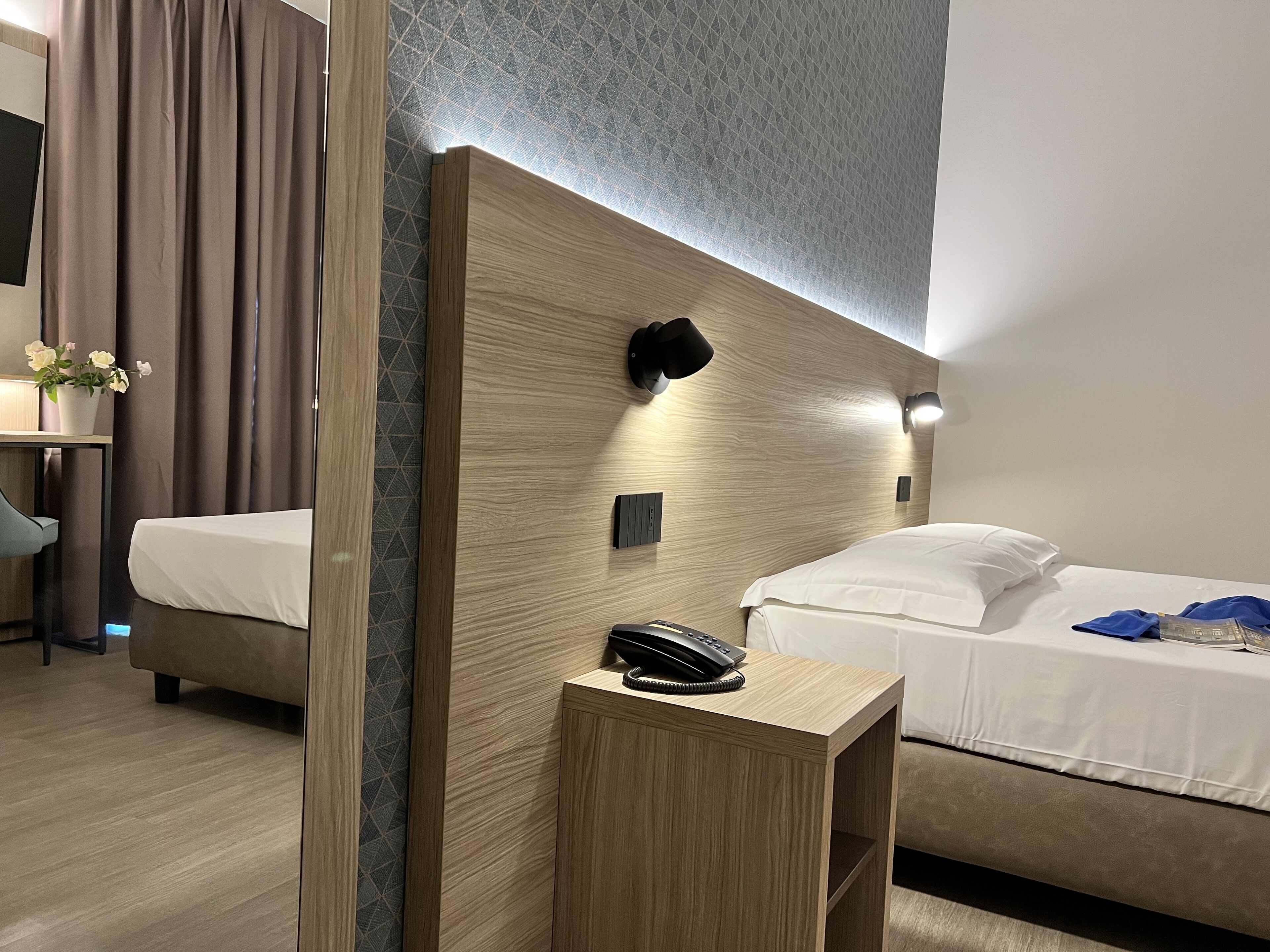 Une chambre d’hôtel avec une tête de lit en bois, une table de chevet avec un téléphone, un lit avec une literie blanche et un bureau avec une chaise.
