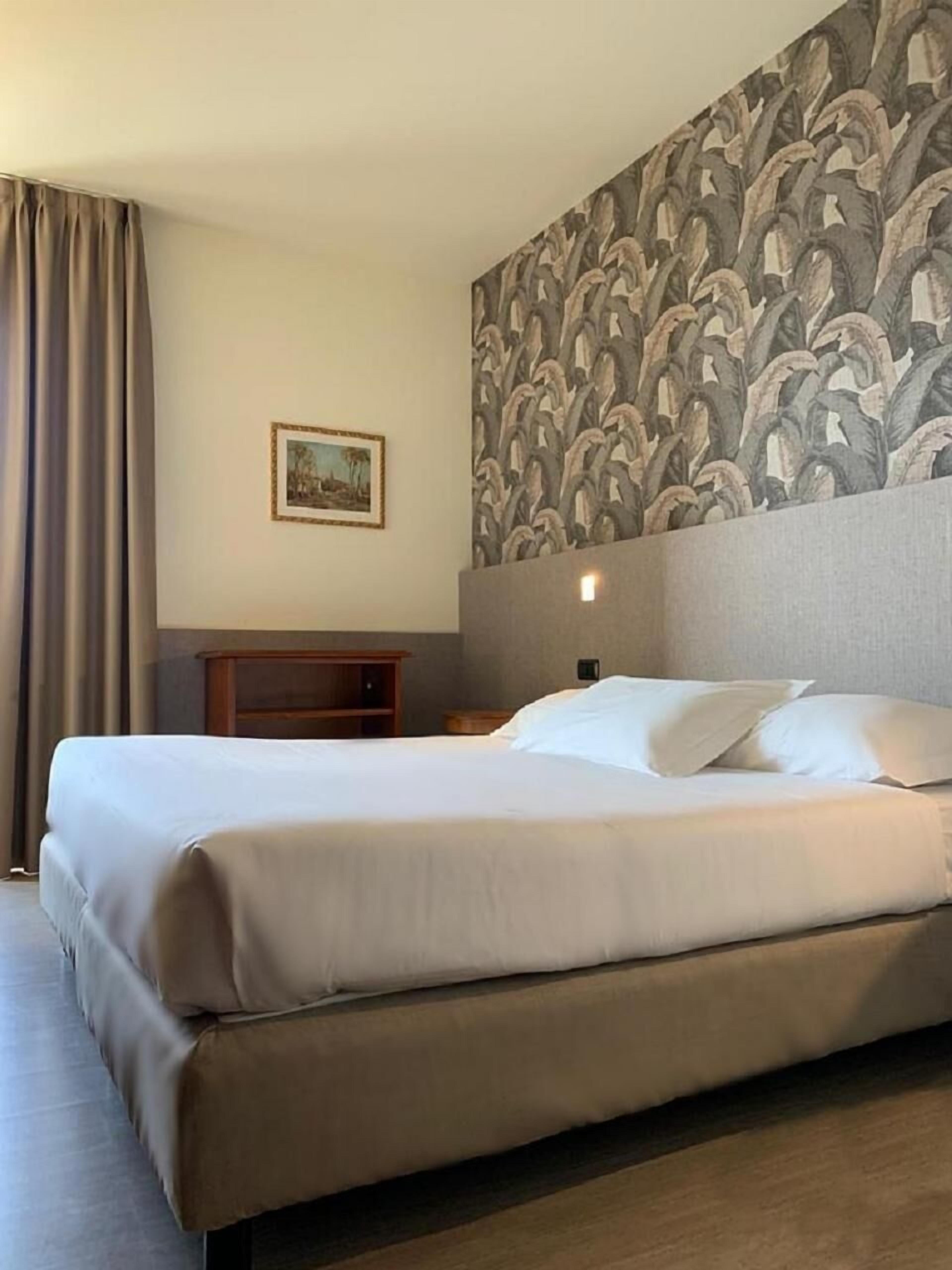 Una habitación de hotel con una cama grande, una mesita de noche, un cuadro enmarcado en la pared y un papel tapiz estampado.