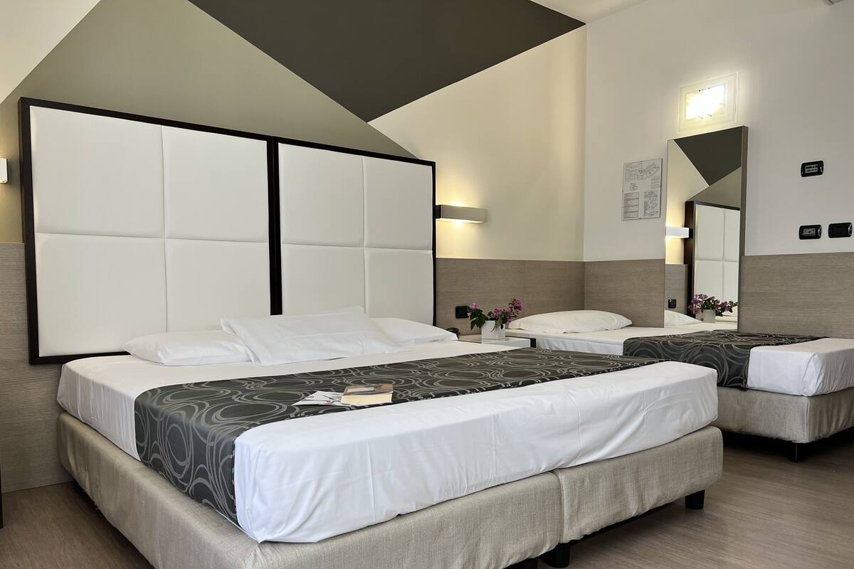 Une chambre d’hôtel avec deux lits, une grande tête de lit rembourrée, une table de chevet ornée d’un vase de fleurs et un miroir au mur.