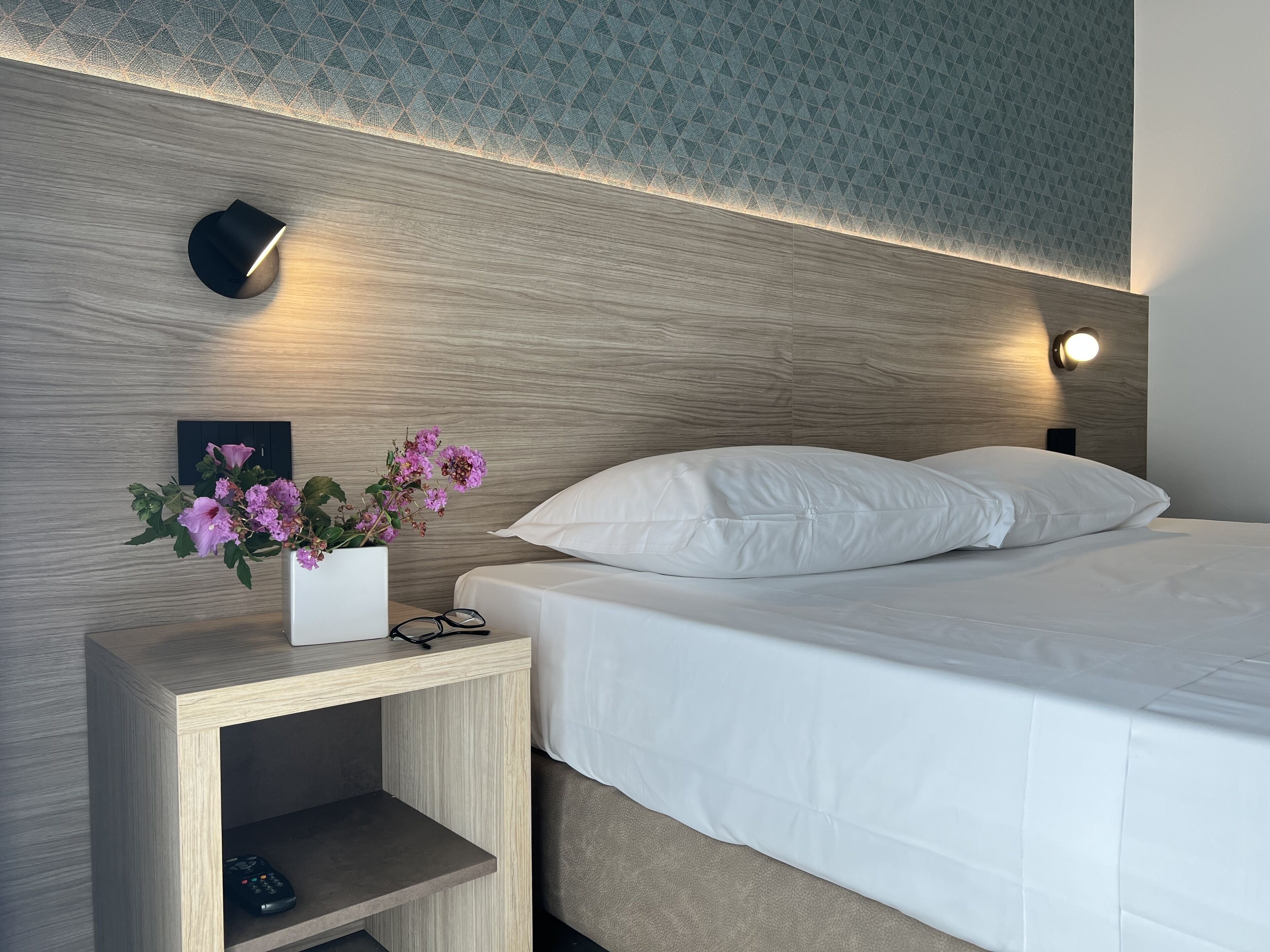 Una cama bien hecha con ropa blanca, una mesita de noche con un florero y dos lámparas fijadas a la pared.
