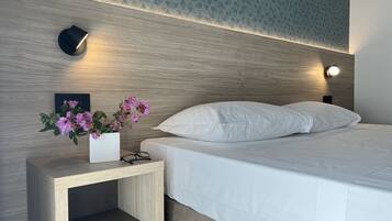 Una cama bien hecha con ropa blanca, una mesita de noche con un florero y dos lámparas fijadas a la pared.