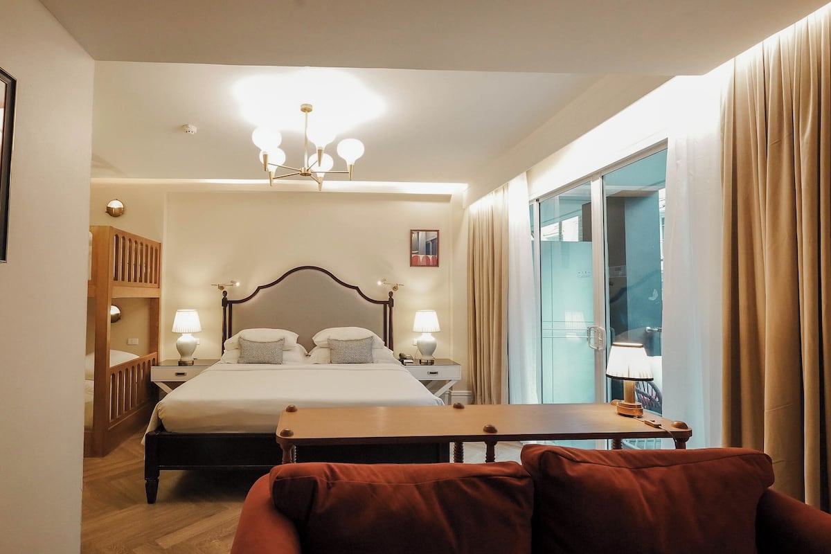 Habitación de hotel con cama, mesitas de noche, escritorio, silla, armario, espejo y ventana con cortinas.