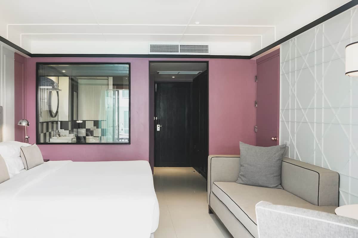Una habitación de hotel moderna con una cama grande, un sofá, un espejo y una puerta que da a otra habitación.