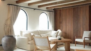 Un salón acogedor con un sofá blanco, una mesa de centro de madera y una silla. Hay dos ventanas grandes con cortinas, una pared de madera y un jarrón grande con ramas.