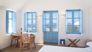 A room with a bed, a table with chairs, a window with blue shutters, and a view of the sea. - Pantelia Suites (Santorini)