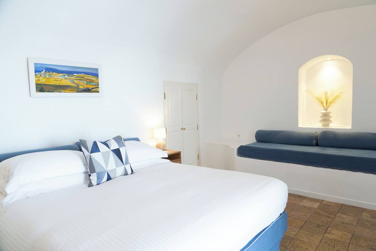 Un dormitorio con cama, un asiento junto a la ventana, una puerta y un cuadro en la pared.