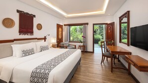 A hotel room with a large bed, a desk, a chair, a TV, and a view of greenery outside.