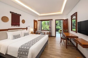 A hotel room with a large bed, a desk, a chair, a TV, and a view of greenery outside.