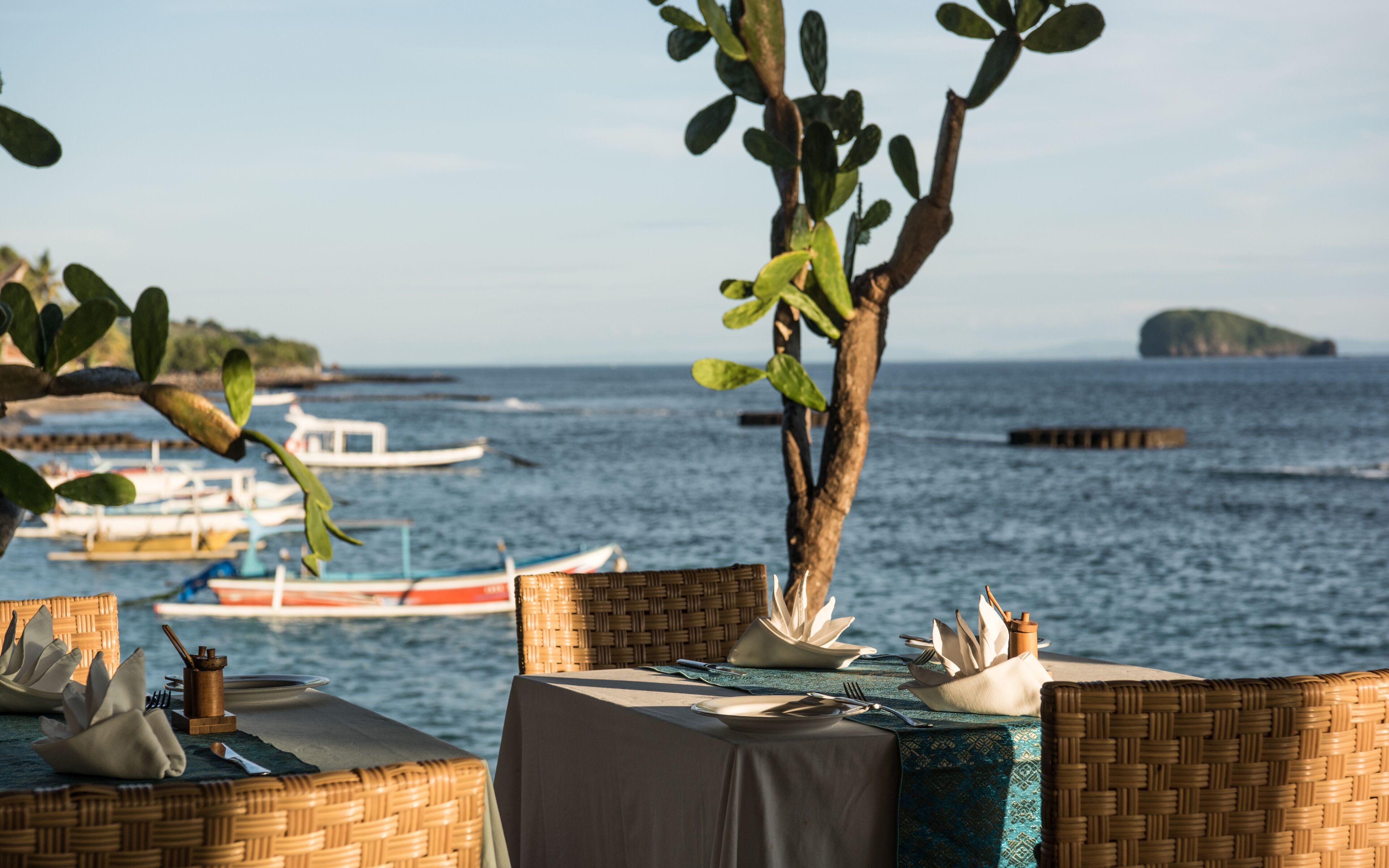 A seaside dining area with wicker furniture and a table set for two, overlooking the sea and boats.