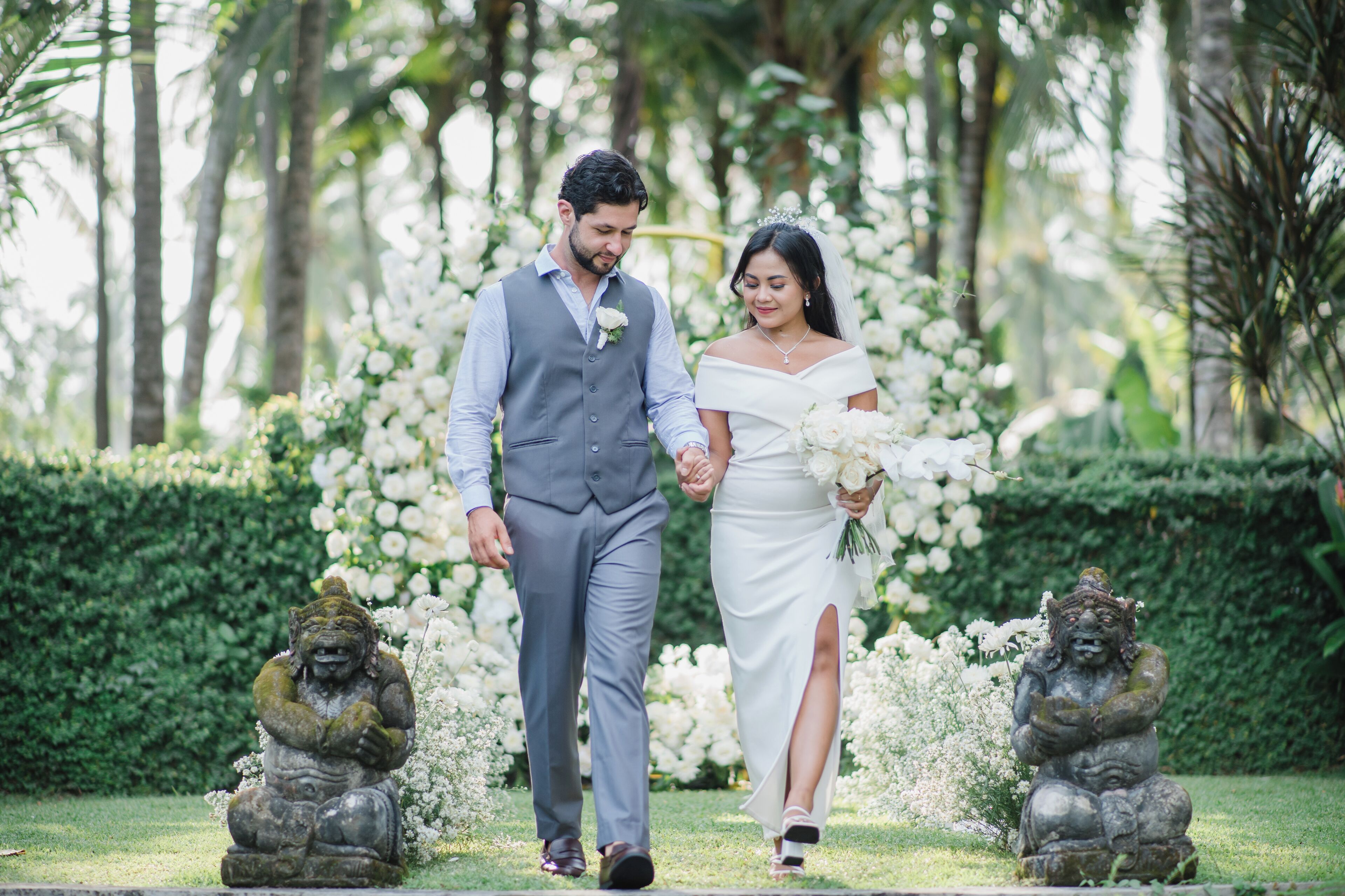 Una pareja caminando de la mano en un jardín con estatuas y flores blancas.
