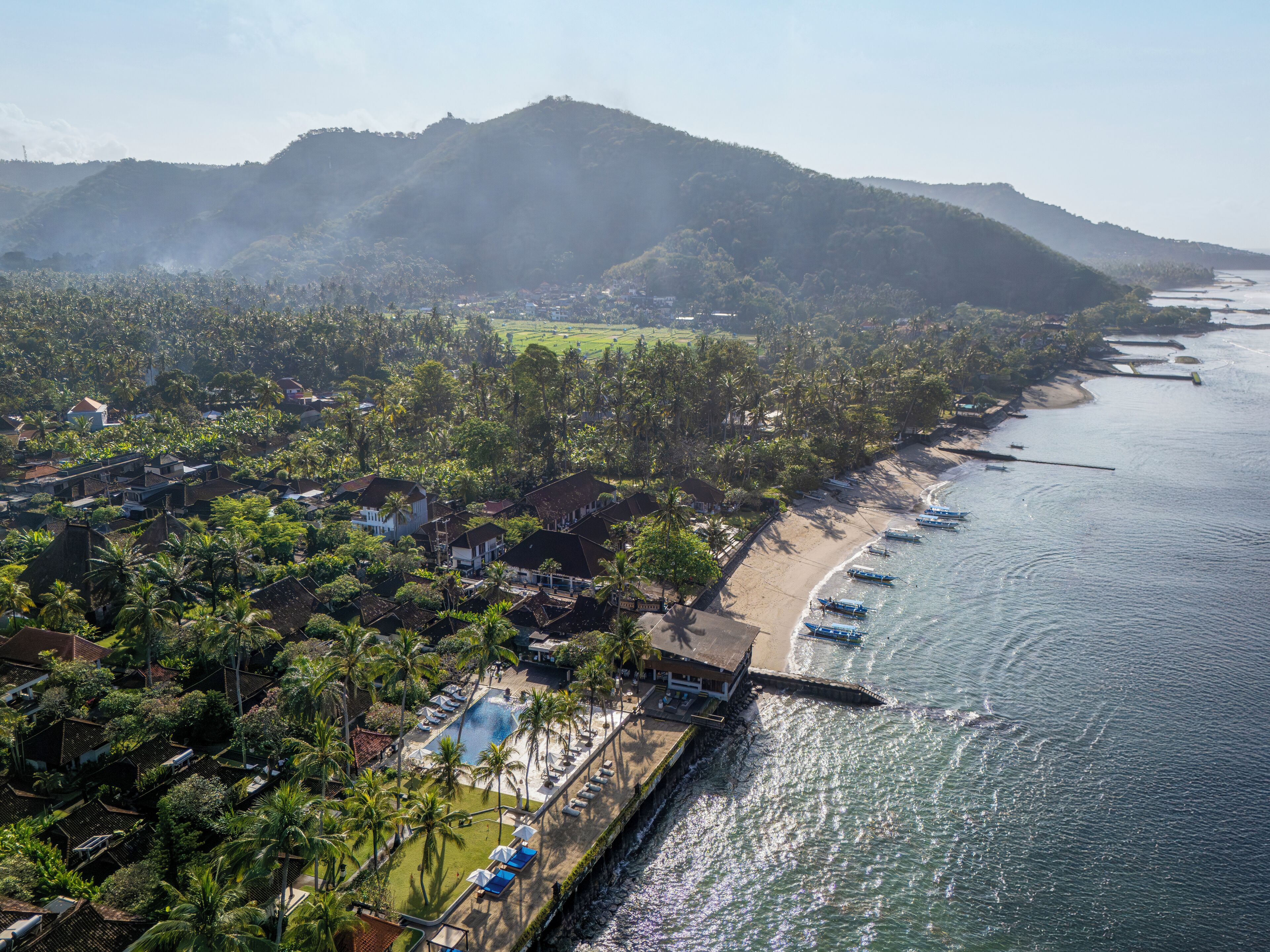 An aerial view of a coastal resort with a beachfront, a swimming pool, and a view of the surrounding hills.