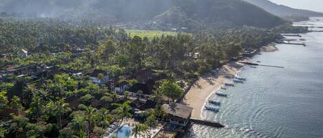 Vista aérea de un complejo turístico en la costa, con playa, piscina y vistas de las colinas circundantes.