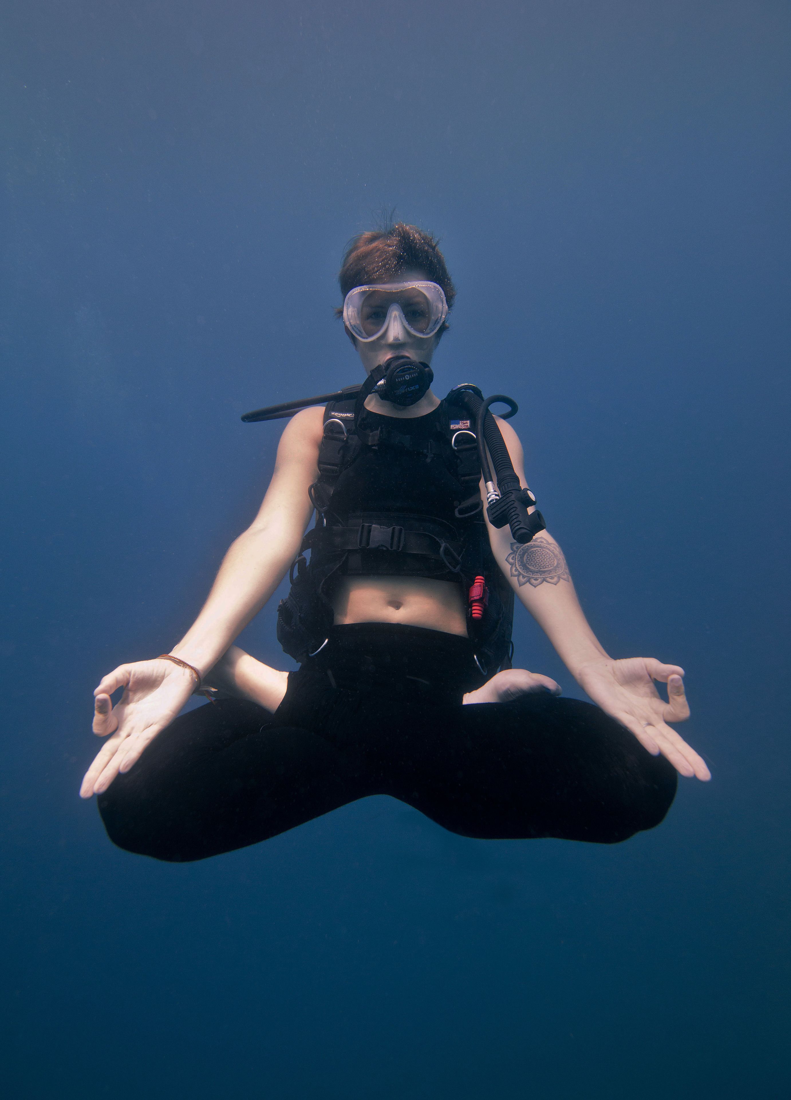A person in a black wetsuit and scuba gear sitting underwater with a clear blue background.