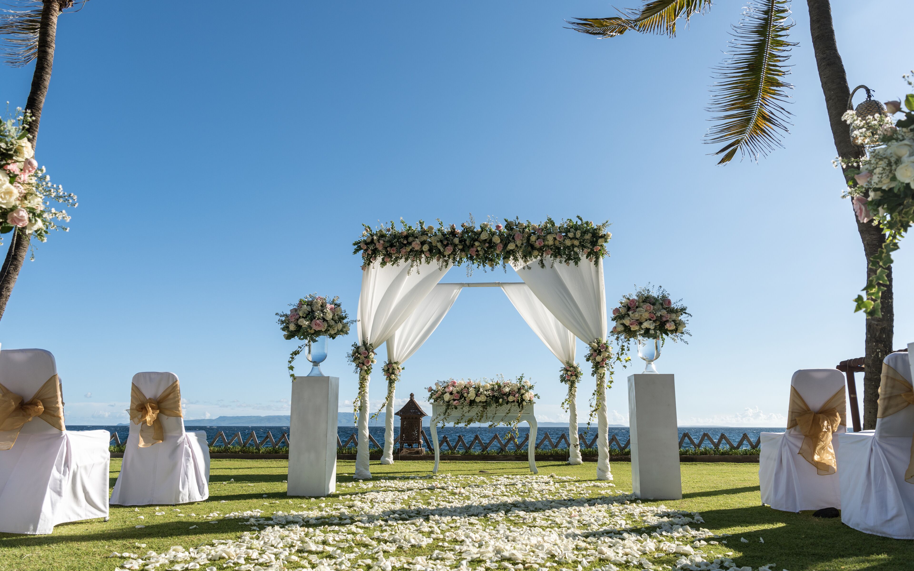 Montaje de ceremonia de boda al aire libre con arcos blancos adornados con telas y arreglos florales, con vista al mar.