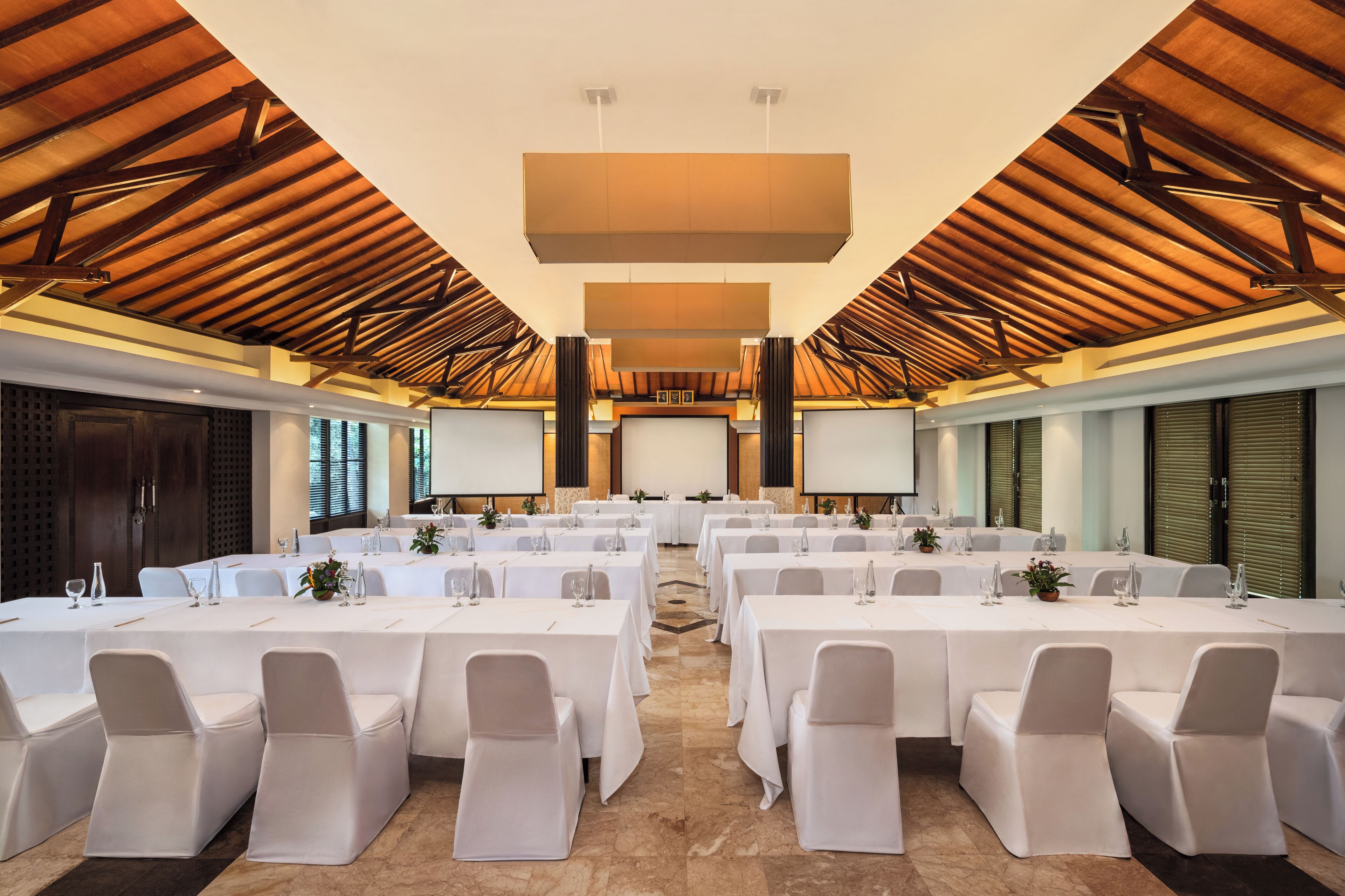 A conference room with multiple tables and chairs, a large screen, and a wooden ceiling structure.