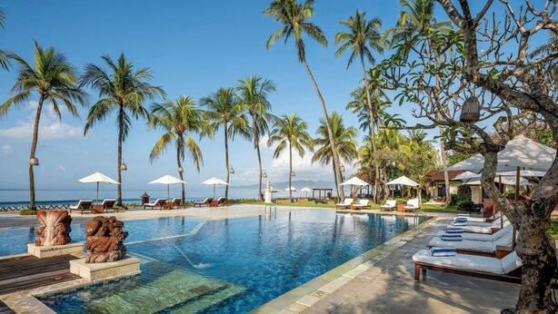 A poolside area with lounge chairs and umbrellas, palm trees, and a clear view of the ocean.