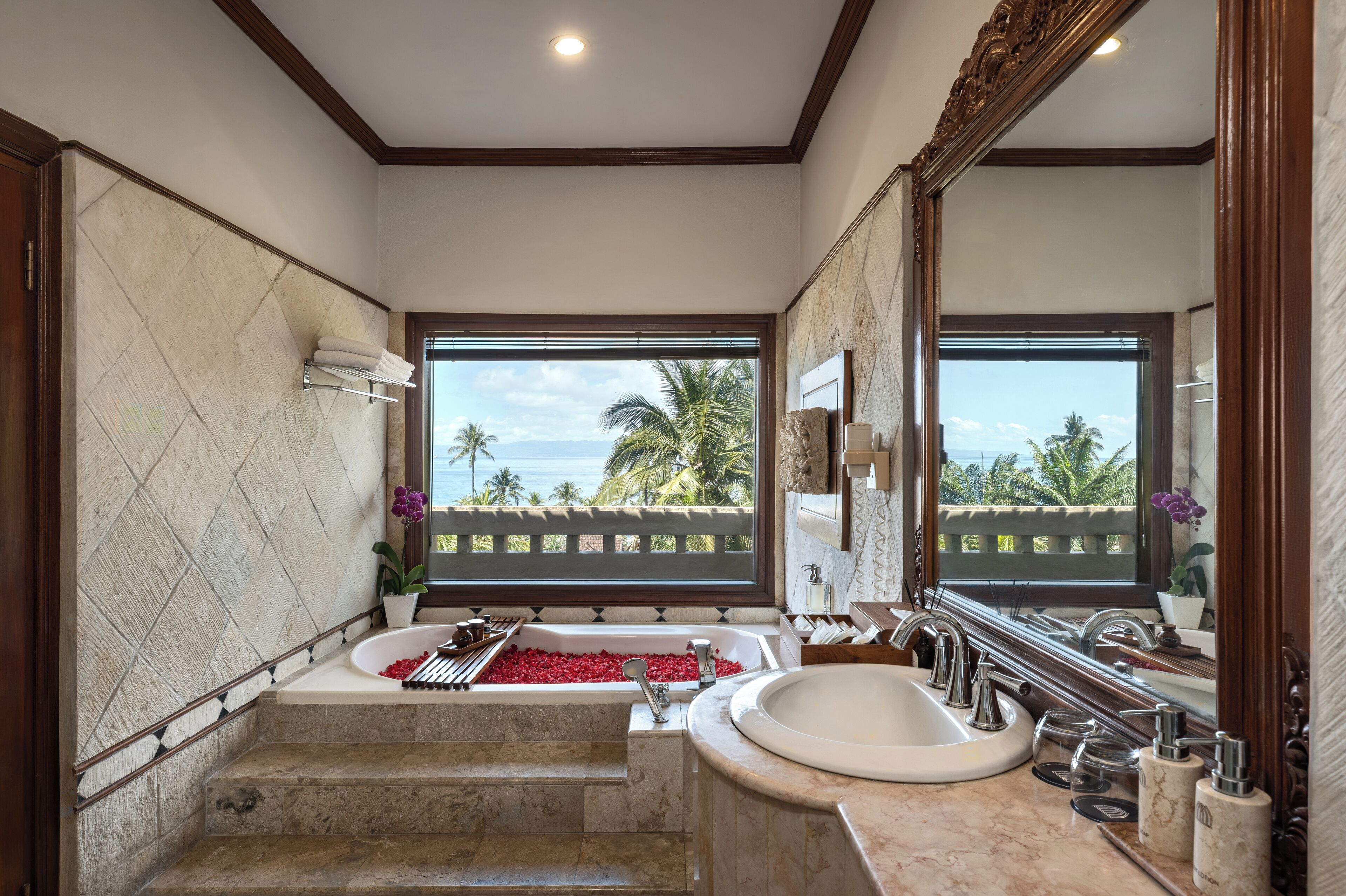 A bathroom with a bathtub, a sink, and a large mirror. There is a view of palm trees and the ocean through the windows.