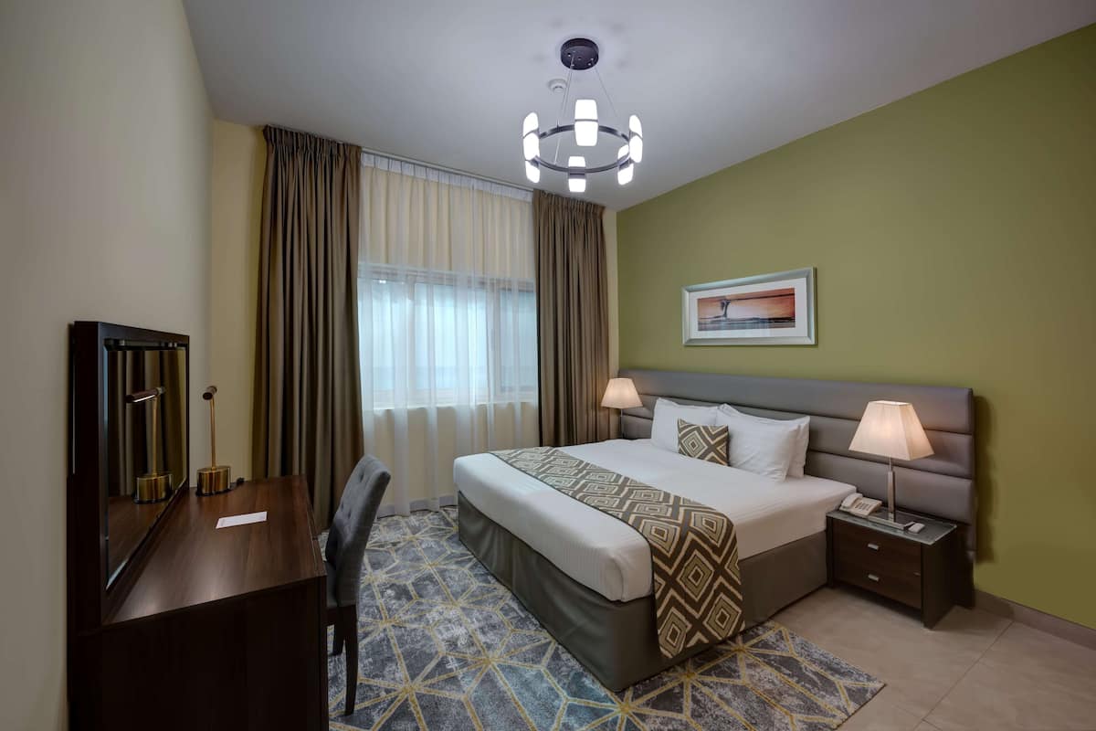 Une chambre d’hôtel avec un lit, des lampes de chevet, un bureau avec une lampe, une chaise, une fenêtre avec des rideaux et un tableau au mur.