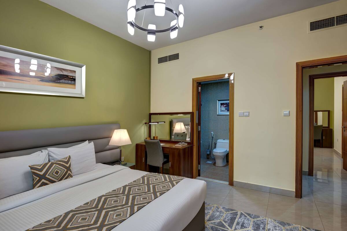 Une chambre d’hôtel avec un grand lit, des tables de chevet, un bureau, une chaise, une lampe de bureau, un miroir, un tableau encadré au mur et une salle de bain visible à travers une porte ouverte.