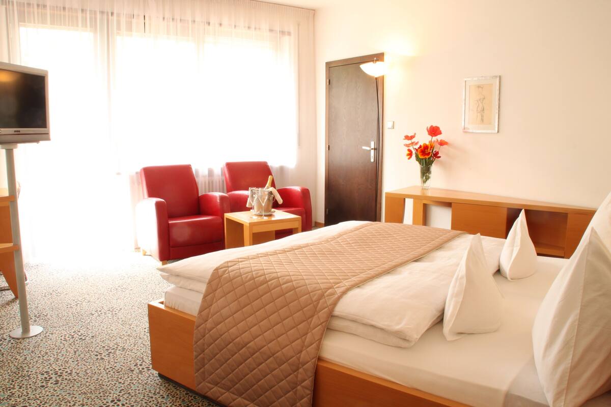 A hotel room with a bed, a red armchair, a small table with a vase of flowers, and a television.