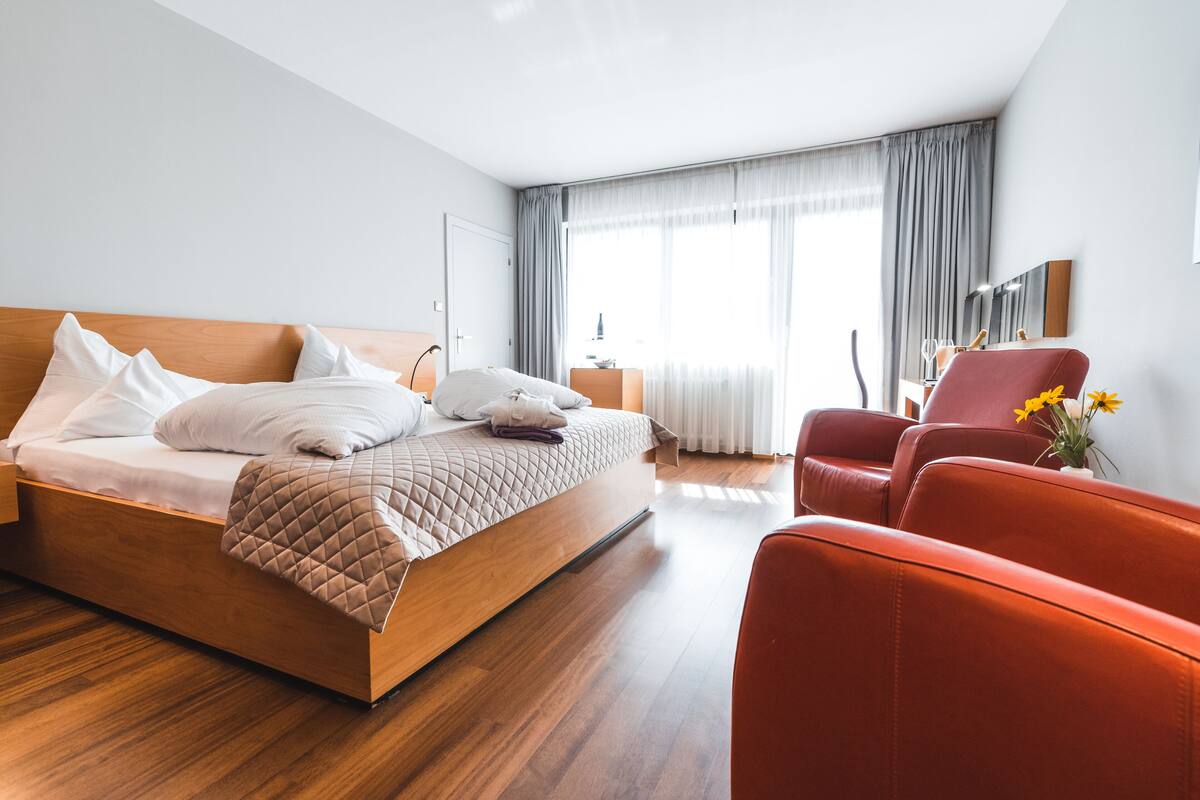 A modern hotel room with a wooden bed, a red armchair, and a large window with curtains.