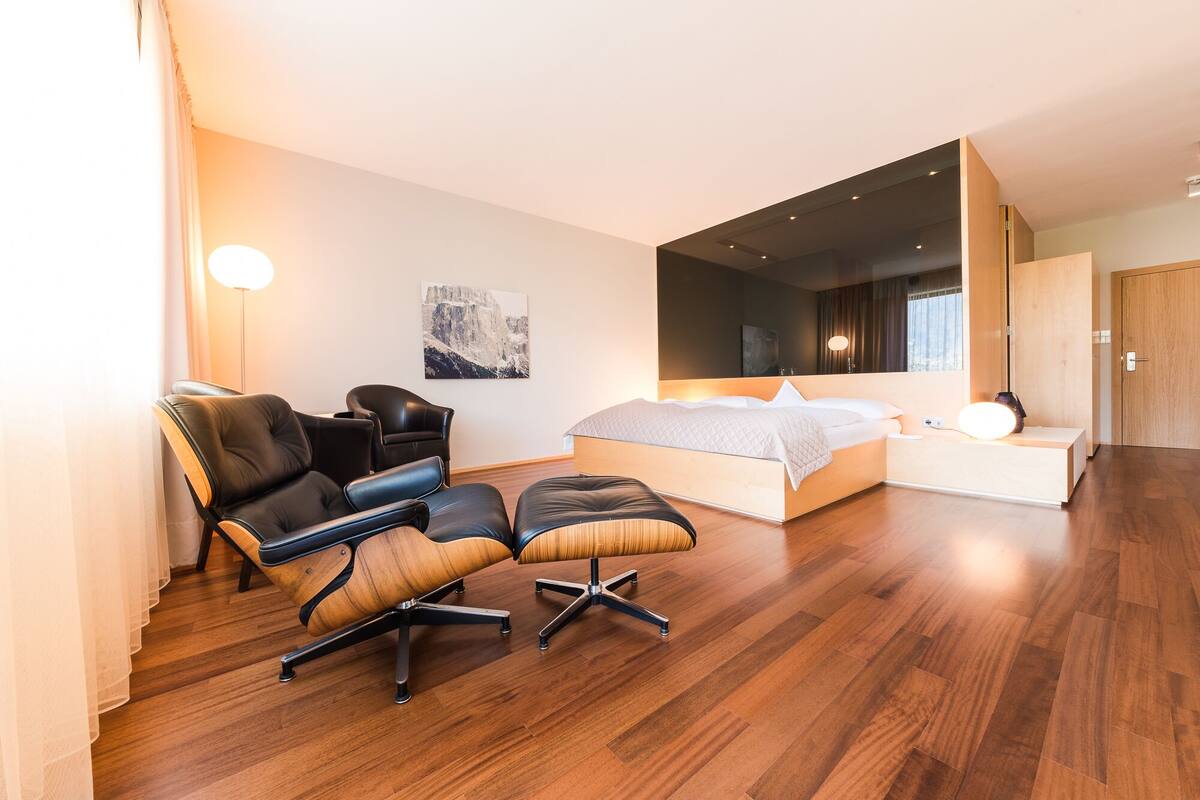 A modern bedroom with a large bed, two black leather chairs, a wooden ottoman, a bedside table with a lamp, and a large window.