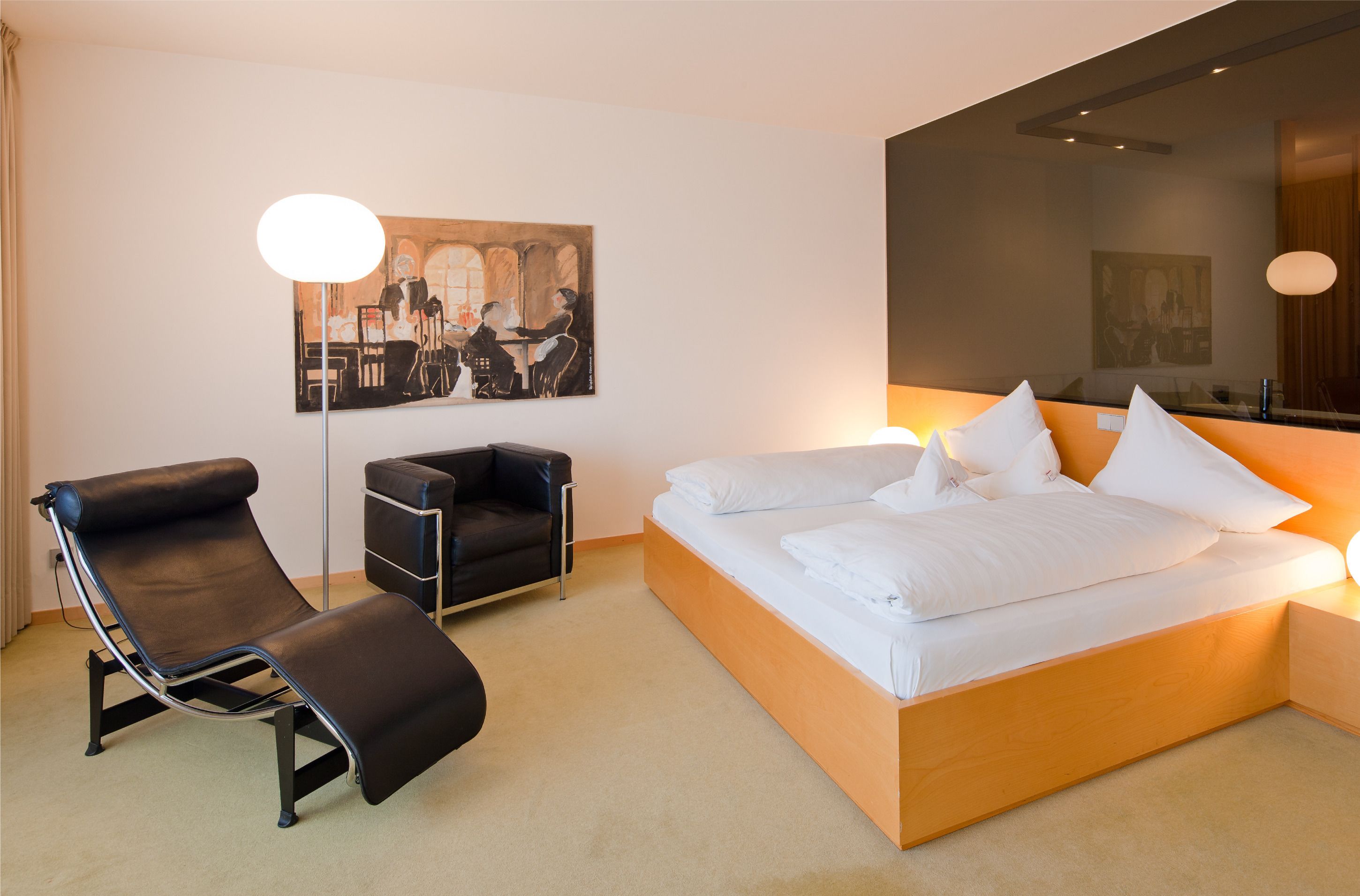 A modern hotel room with a single bed, a reclining chair, a small desk, and a large mirror.