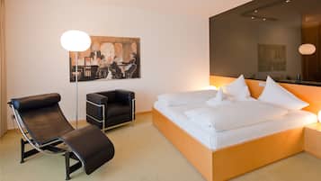 A modern hotel room with a single bed, a reclining chair, a small desk, and a large mirror.