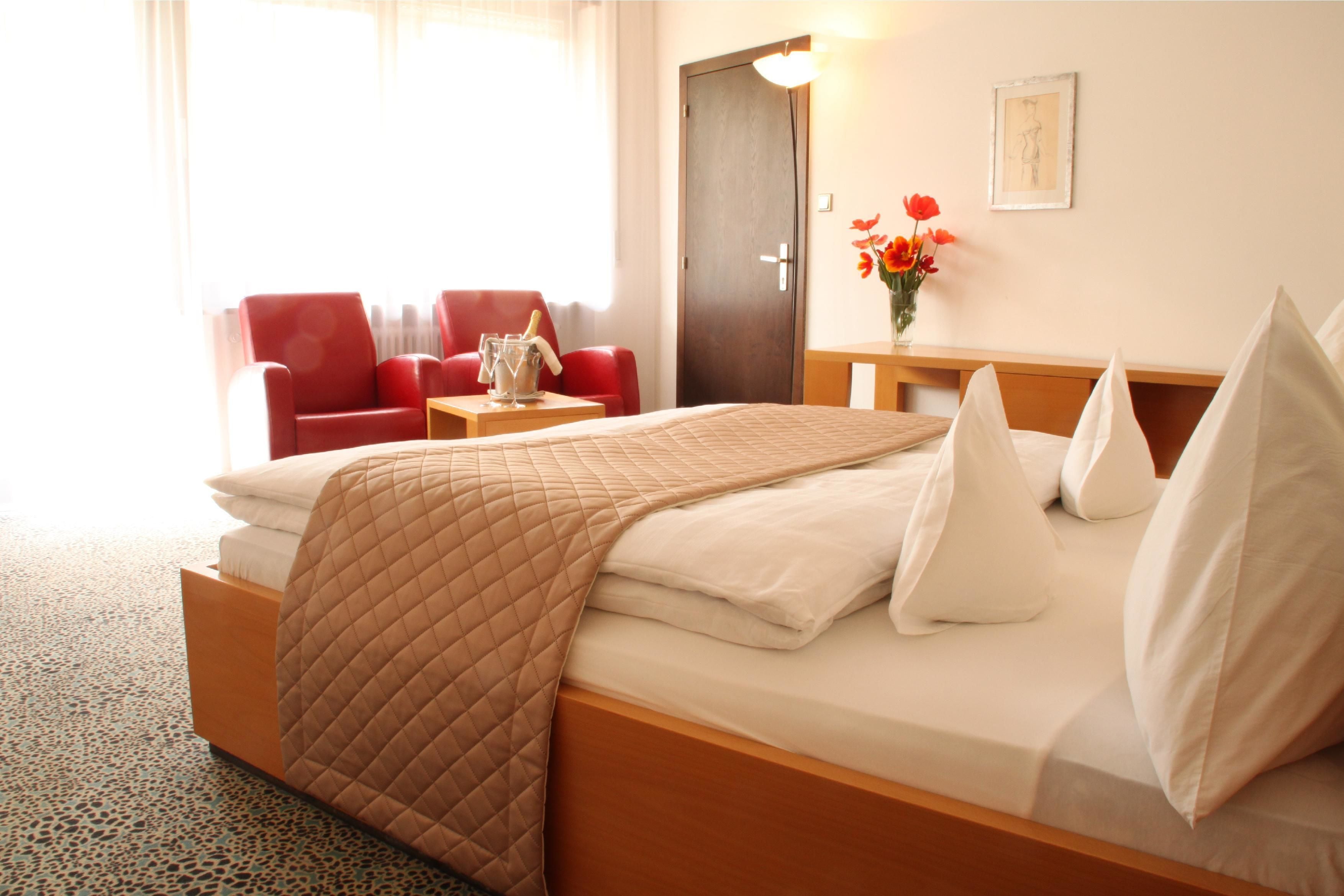 Una habitación de hotel con una cama, un sillón rojo, una mesita con un florero y una puerta de madera.