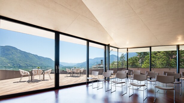 A spacious room with large windows offering a mountain view, a wooden deck, and modern seating. - Hotel Marlena (Marlengo)