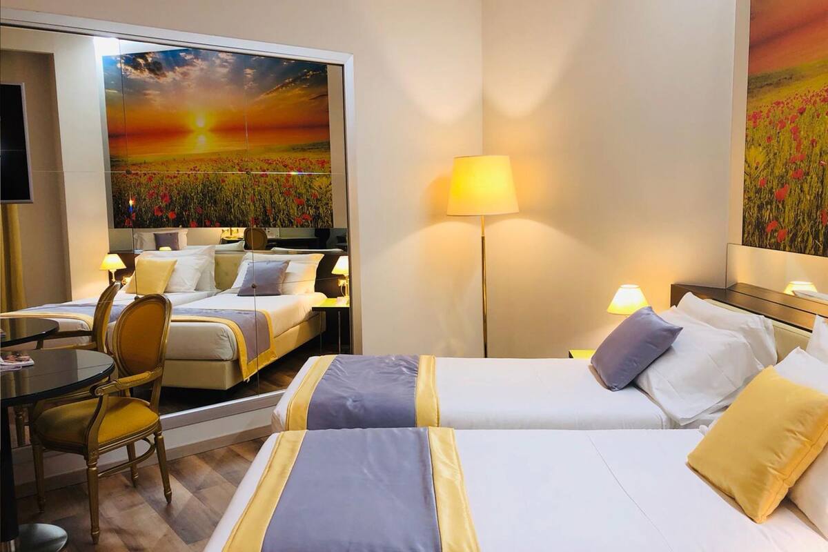 A hotel room with two beds, a desk, a chair, and a painting of a sunset.