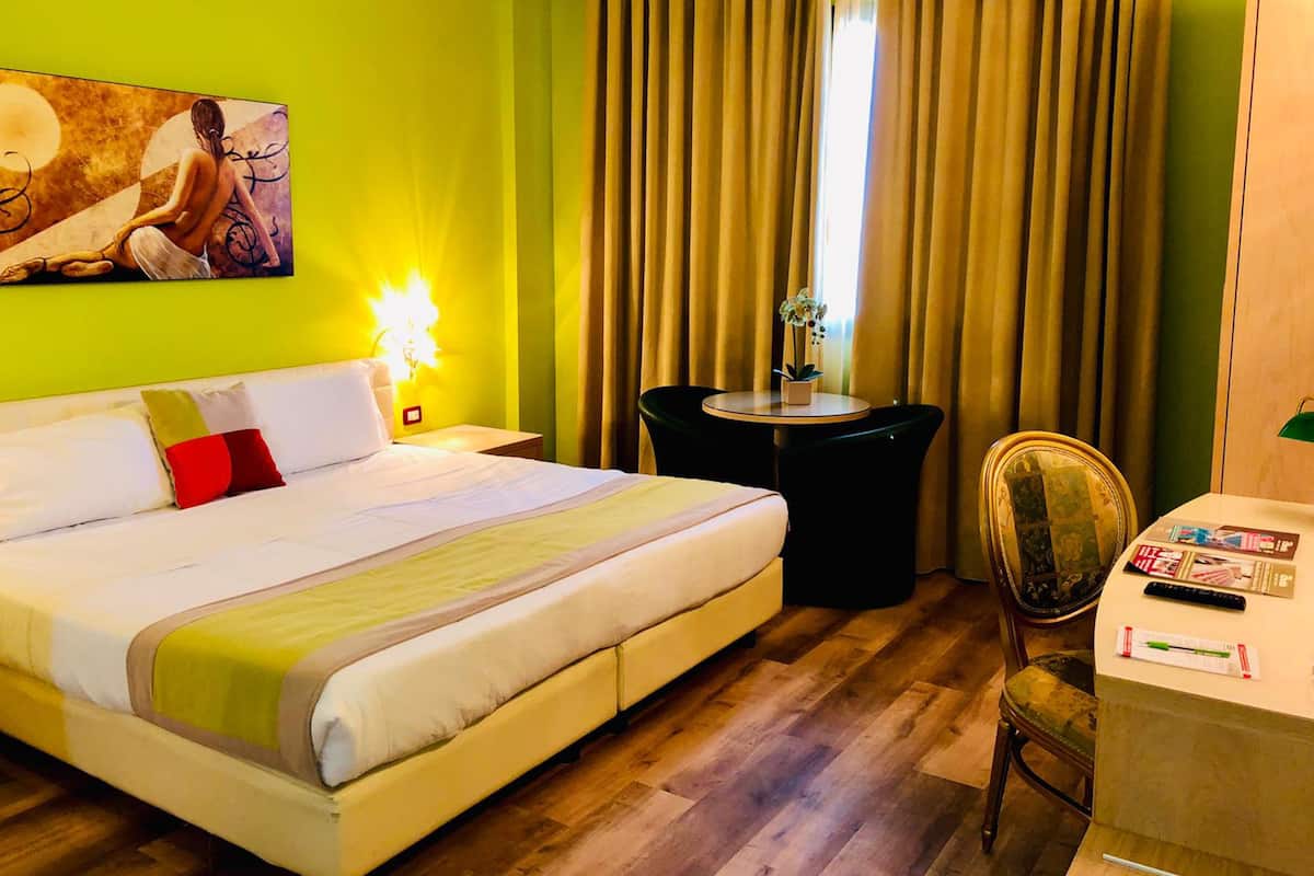 A hotel room with a bed, a desk, a chair, a small table with a plant, and a painting on the wall.