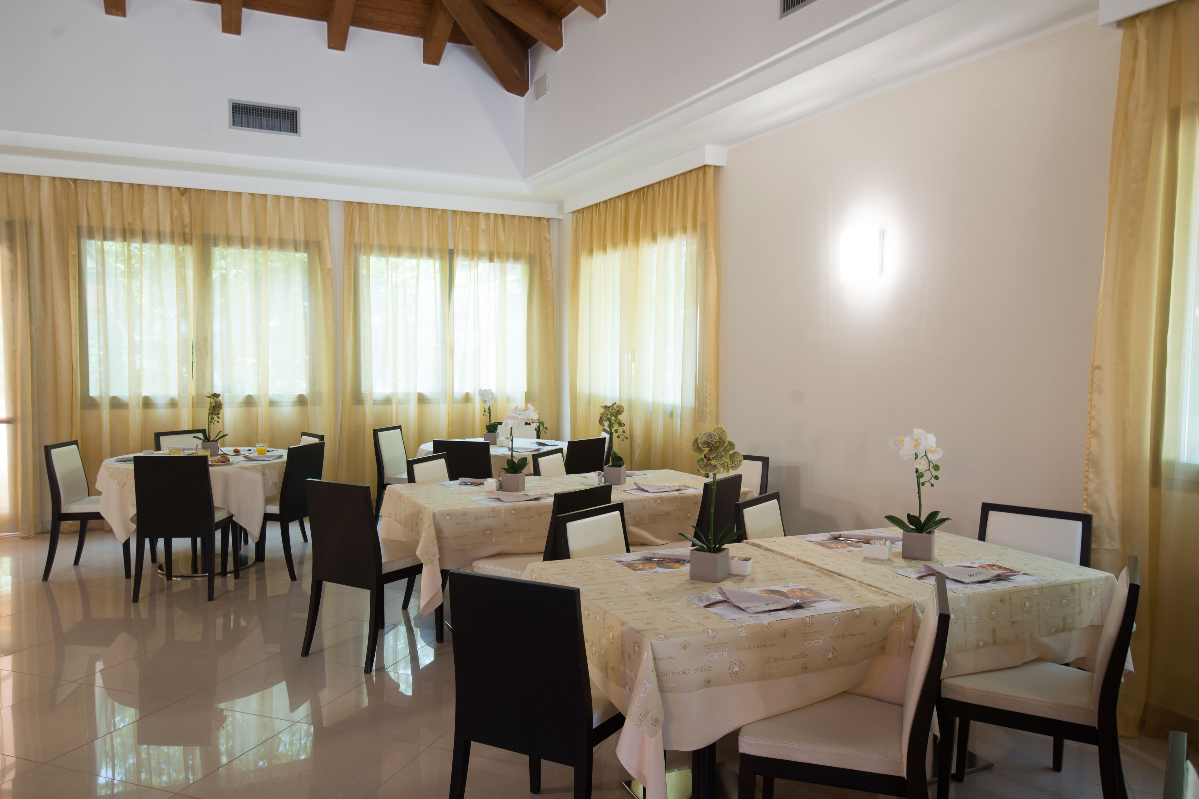 Comedor con varias mesas y sillas, dispuesto en una sala amplia con ventanas grandes y cortinas.