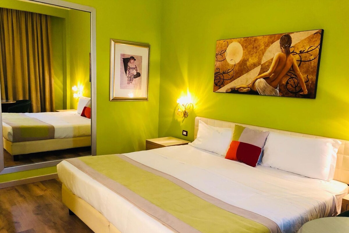 A hotel room with a large bed, a mirror, a framed picture, and a wall painting.