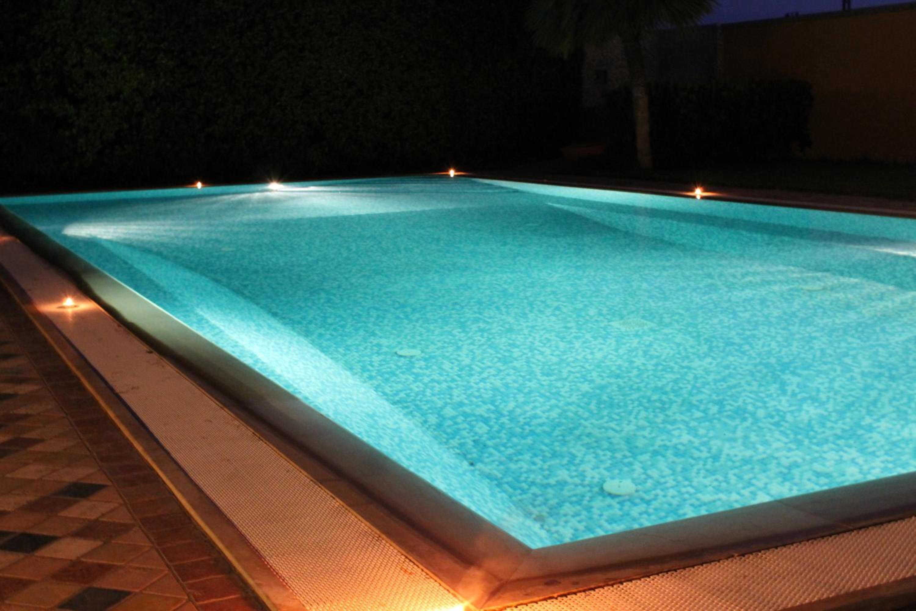 Una piscina rettangolare con acqua cristallina, circondata da un solarium piastrellato e illuminata da luci.