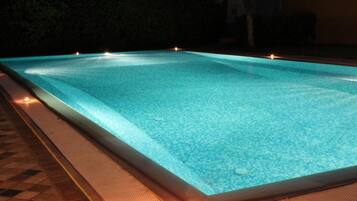 Una piscina rettangolare con acqua cristallina, circondata da un solarium piastrellato e illuminata da luci.