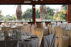 A restaurant with tables set for dining, large windows with a view of palm trees and greenery.