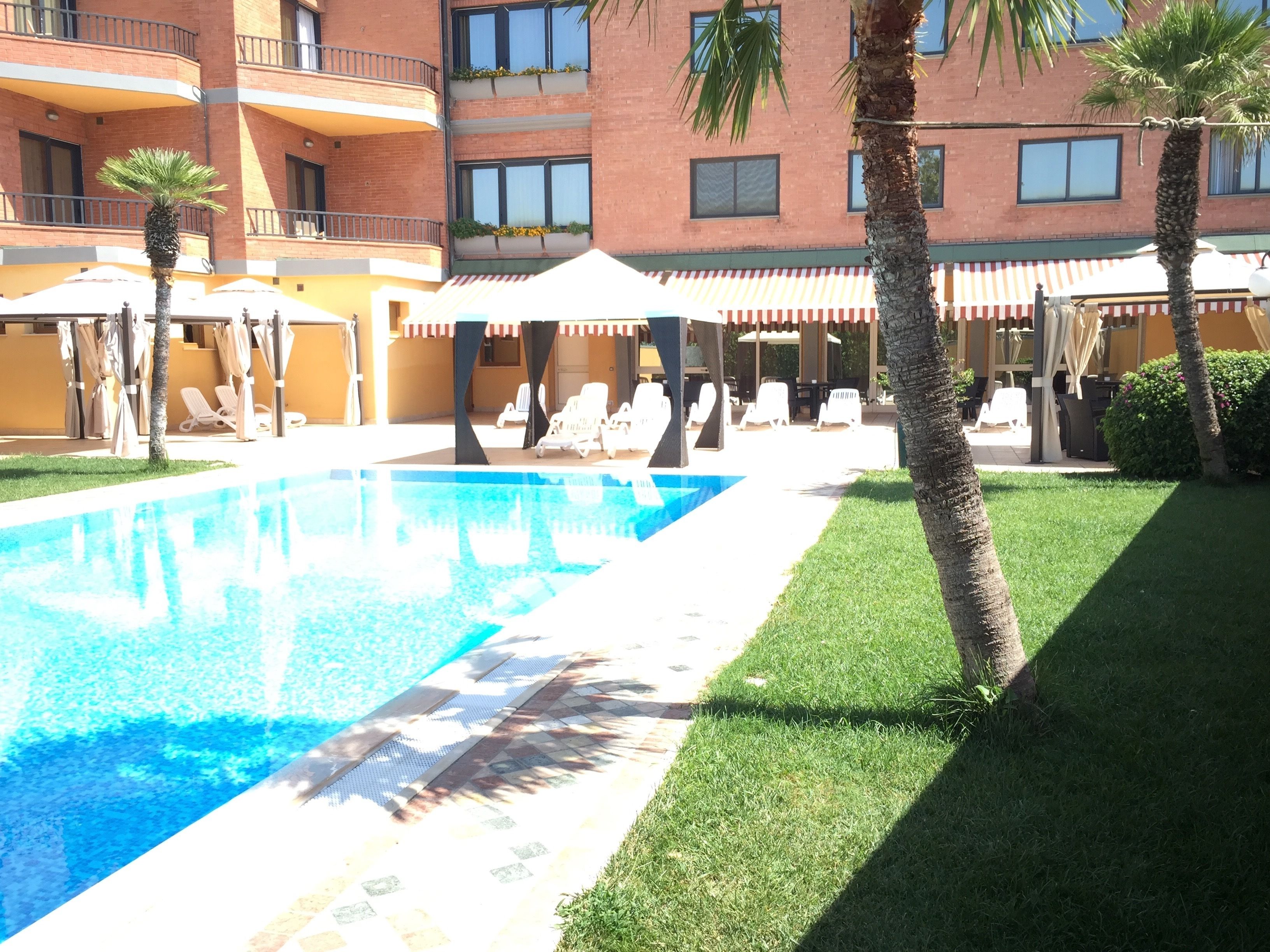 A swimming pool with lounge chairs and umbrellas, surrounded by palm trees and a building with balconies.