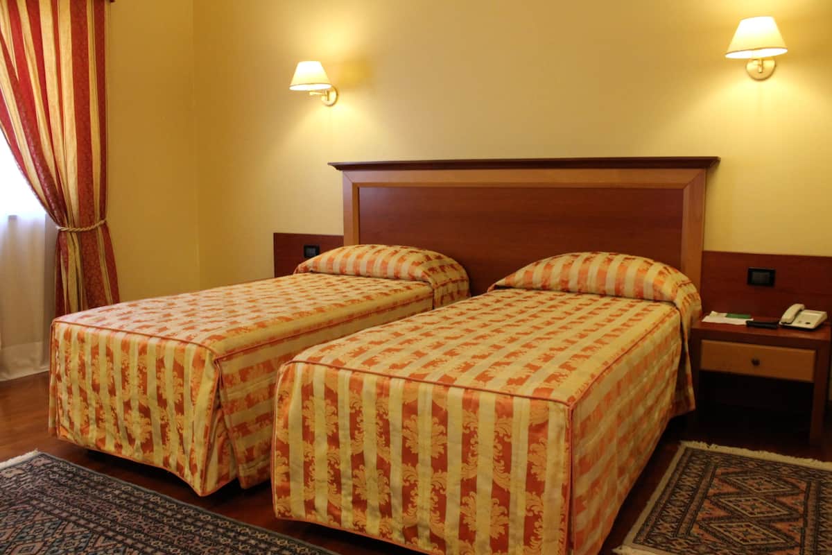 Hotellihuone, jossa on kaksi erillistä sänkyä, yöpöytä, jossa on puhelin, ja seinälle asennettu lamppu.