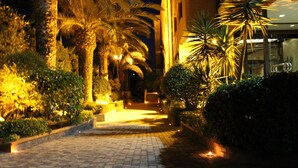 A paved pathway flanked by palm trees and shrubs, leading to a building entrance.