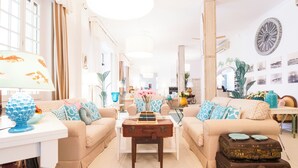 A well-lit living room with two beige sofas, a coffee table, and a small table with a lamp. There are decorative pillows and a vase with flowers on the sofas. A suitcase is placed on the floor next to the sofas. - La Pineta Al Mare (Forte dei Marmi)