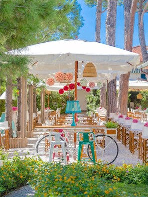 An outdoor dining area with tables and chairs under a large umbrella, surrounded by trees and plants. - La Pineta Al Mare (Forte dei Marmi)