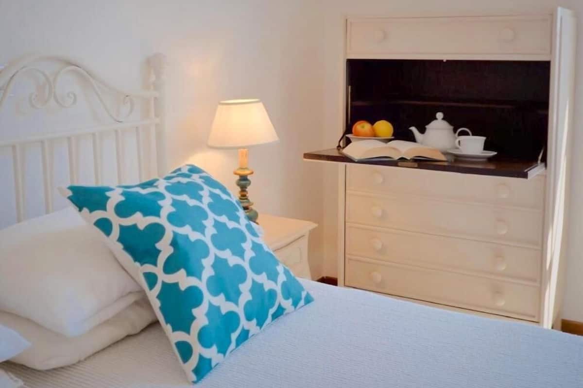 Un lit bien fait, orné d’un oreiller à motifs blancs et bleus, une lampe de chevet et une commode avec un miroir et une étagère où sont posés un livre, une théière et une tasse.