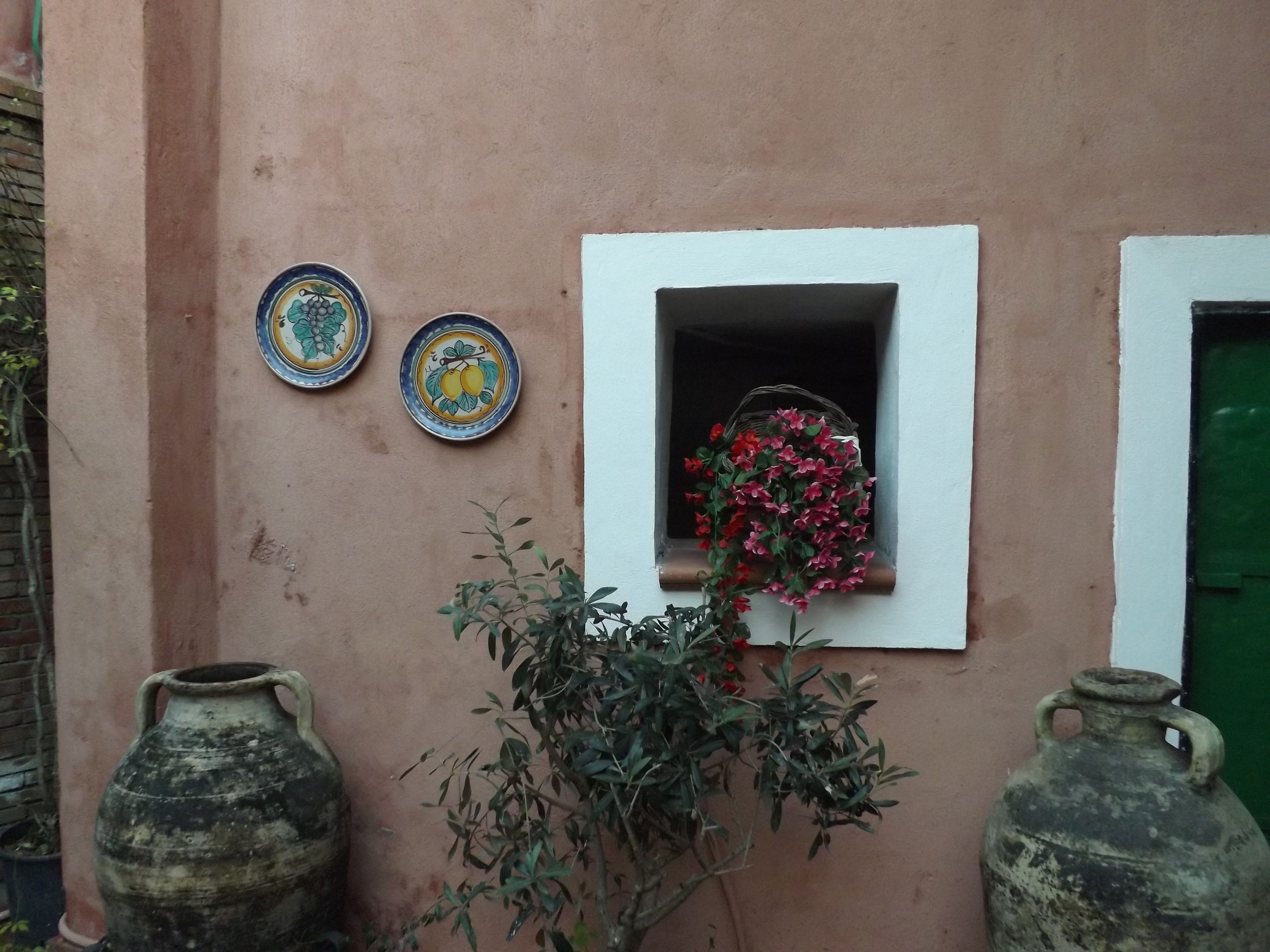 A rustic building with a small window adorned with artificial flowers, two ceramic plates with floral designs, and two large ceramic pots.